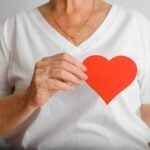 World stroke day, heart disease concept. Close-up of elderly woman holding red paper heart with wrinkled hands on background of white t-shirts. Selective focus on the heart