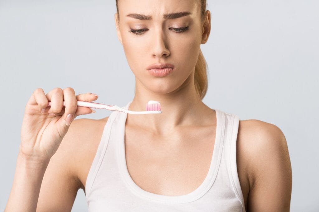 Displeased Girl Looking At Toothbrush Cleaning Teeth Over Gray Background